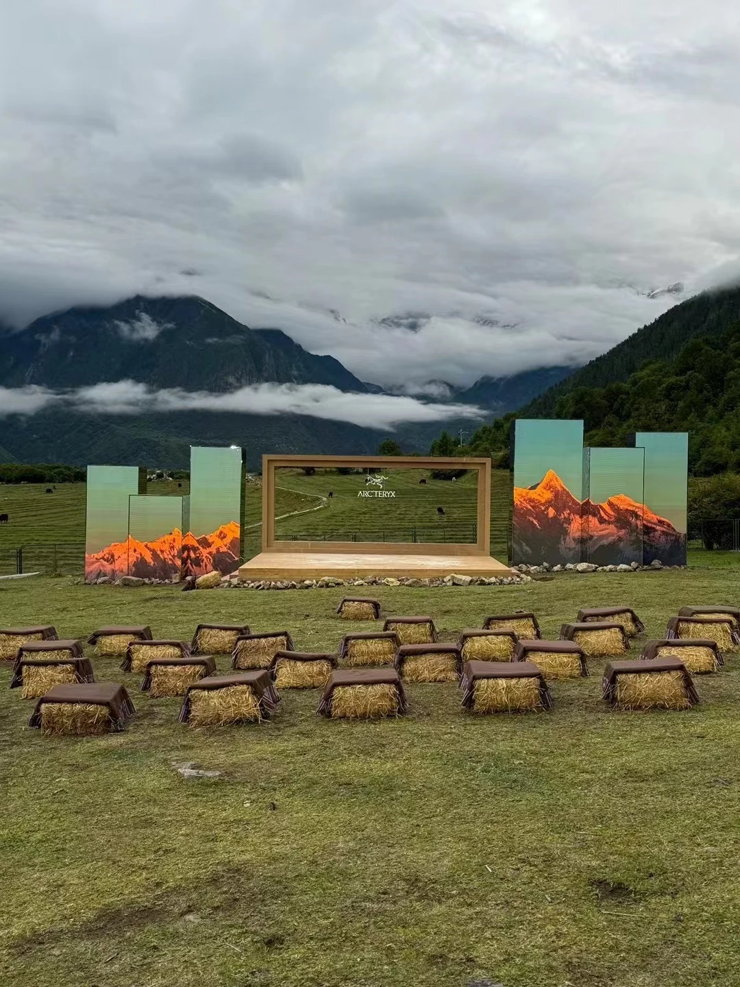 雪山和云层下的完整会场，草捆座椅面向木框舞台和两侧山景屏